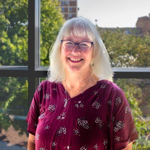 Smiling woman with silver hair and glasses wearing a burgundy floral blouse, standing outdoors on a sunny day.
