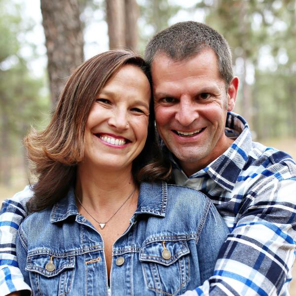 Smiling couple with arms around each other outdoors in a wooded area, both wearing denim jackets and plaid shirt.