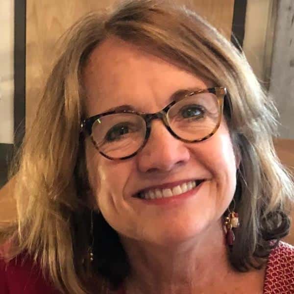Smiling woman with short, light brown hair and tortoiseshell glasses, wearing a red top and dangly earrings.