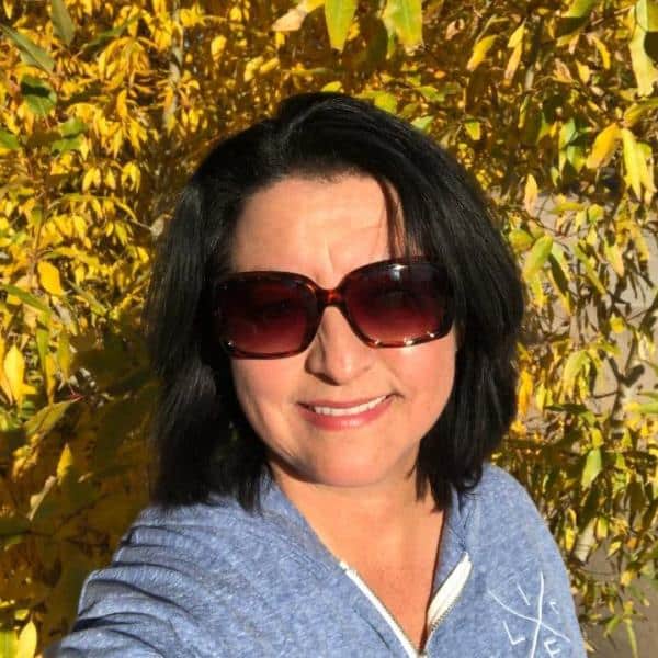 Woman with shoulder-length black hair wearing large red sunglasses, smiling outdoors among yellow autumn leaves, in a blue zip-up jacket