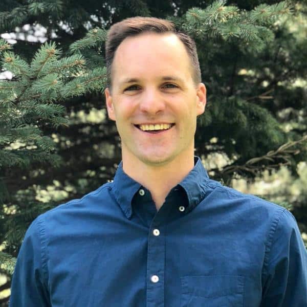 Smiling man in a blue button-down shirt standing outdoors with evergreen trees in the background.