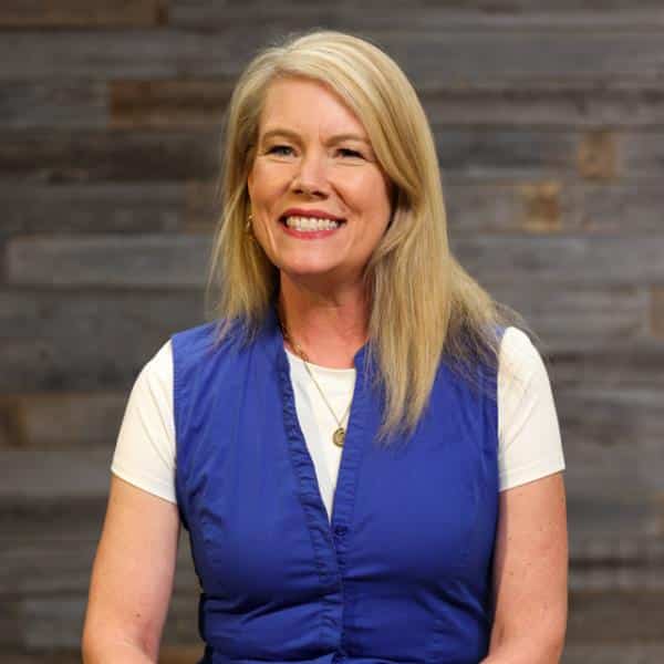 Smiling blonde woman wearing a blue vest over a white shirt, standing against a wood-paneled background.