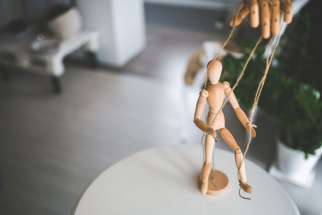 Wooden marionette puppet manipulated by strings over a white round table in a bright indoor room.